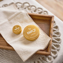 Load image into Gallery viewer, Two gold containers of &#39;navy&#39; hand balm on a wooden tray with a beige cloth and textured surface.