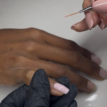 Load image into Gallery viewer, A close-up of a manicure in progress, showing a hand with a light pink nail being painted by another hand holding a fine nail brush, with a black-gloved hand supporting the client's hand on a white surface.