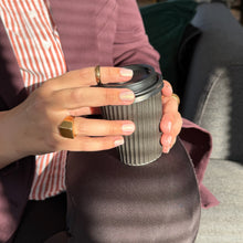 Load image into Gallery viewer, A close-up of a person holding a black paper coffee cup with a lid, wearing a mauve cardigan over a red and white striped shirt, with gold rings on their fingers.