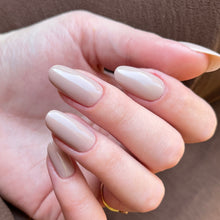 Charger l'image dans la galerie, A close-up of a hand with long, almond-shaped nails painted in a glossy, warm nude beige shade, resting against a brown textured background.