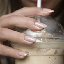 Charger l'image dans la galerie, Close-up of a hand with a glossy nude-pink manicure drinking from a clear plastic cup with a white straw and dome lid, filled with an iced coffee or latte.