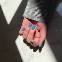 Load image into Gallery viewer, A close-up of a hand with medium blue manicured nails, wearing a black and gray plaid blazer sleeve, resting on a white marble surface with dramatic natural sunlight and shadows.