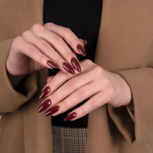 Load image into Gallery viewer, A close-up of a person's hands with long, almond-shaped nails painted in a deep burgundy red, styled with a black top and a brown plaid skirt under a camel-colored coat.