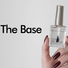 Load image into Gallery viewer, A hand with dark burgundy manicured nails holds a bottle of nail polish against a light gray background. The bottle is labeled 'BIO SCULPTURE THE BASE' and has a metallic silver cap.