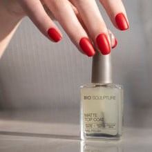 Load image into Gallery viewer, A hand with glossy red manicured nails resting above a bottle of Bio Sculpture Matte Top Coat nail polish on a reflective surface.